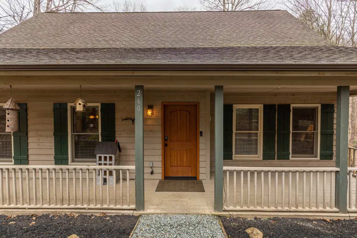 The image shows the inviting exterior of the property, featuring the front door, a welcoming porch area with window shutters, and attractive bird feeders. The setting highlights opportunities to gather on the porch, enjoy nature views, and relax with a book while watching wildlife, all framed by the changing mountain seasons.