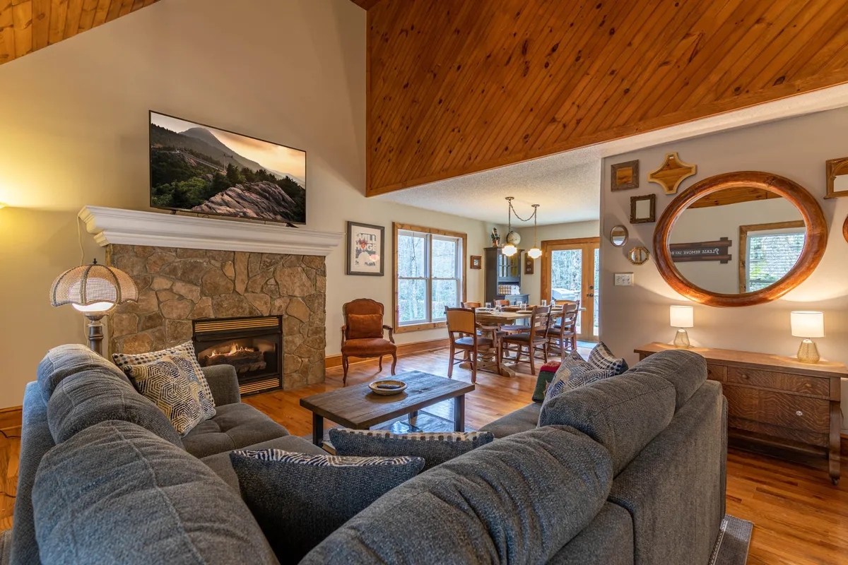 The image showcases a beautifully renovated cabin living and dining area, featuring a stunning stone fireplace as the centerpiece, a large flat-screen TV mounted above, a rustic wooden dining table set for gatherings, and a warm, inviting ambiance enhanced by natural wood details and a decorative mirror. The space is bathed in soft, cozy lighting, creating a perfect environment for relaxation and connection.