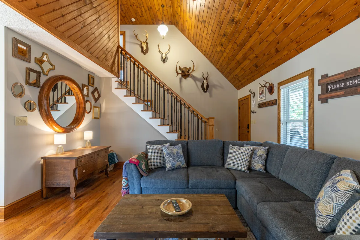 A warm and inviting living room with a sofa facing a coffee table, a wooden staircase ascending in the background, and decorative mirrors and wall-mounted trophies adding charm. The open space is ideal for relaxing after a day exploring with your pet, with ample room for dogs to lounge nearby.
