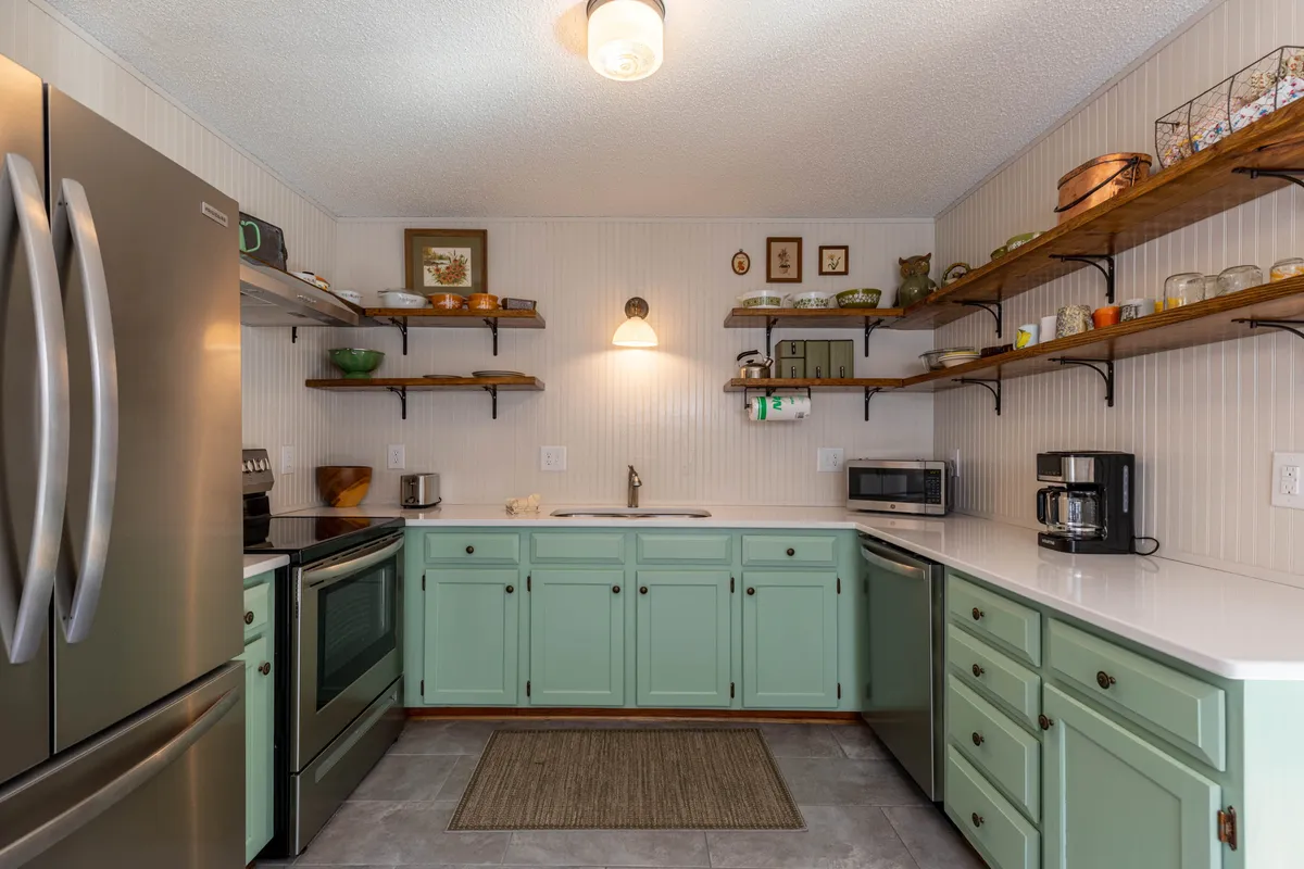 This image features a bright kitchen with mint green cabinetry, open shelving adorned with decorative items, stainless steel appliances, and a subtly textured countertop. The space feels cozy and inviting, with ample light and a layout that encourages cooking and gathering.