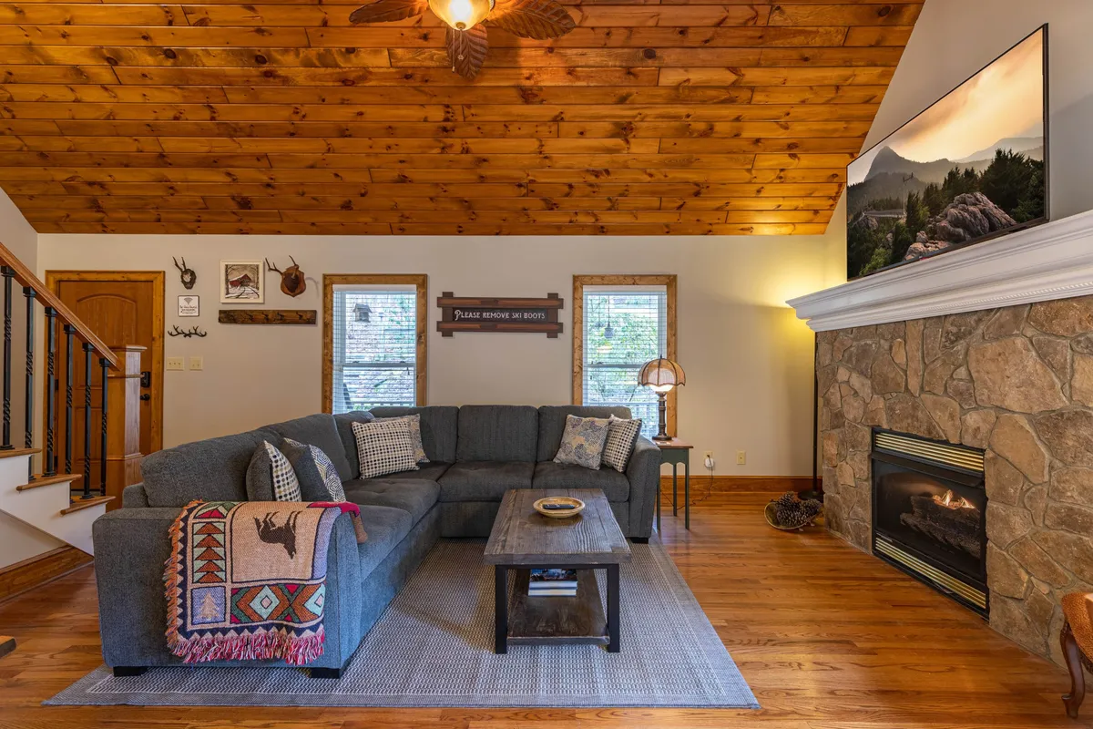 A spacious interior shot with a large sectional sofa facing a stone fireplace, under a wooden tongue-and-groove ceiling. The space includes decorative wall art and is infused with soft, natural light, creating a relaxing, communal environment ideal for lounging or celebrating milestones.