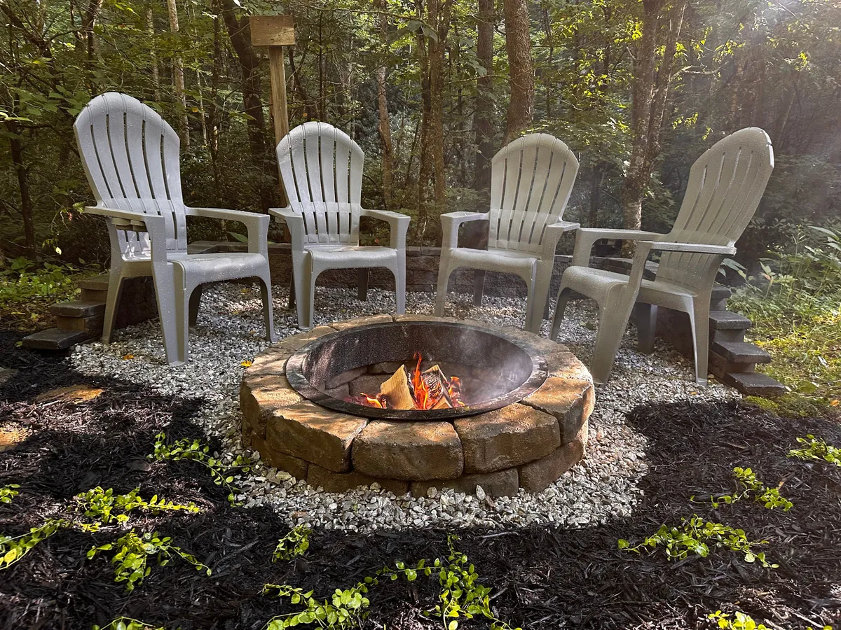 An exterior view of the fire pit area, surrounded by comfortable chairs and lush greenery, ideal for family gatherings, campfire storytelling, making s'mores, and stargazing beneath the mountain sky. The scene is set for evening socialization and shared meals outdoors.