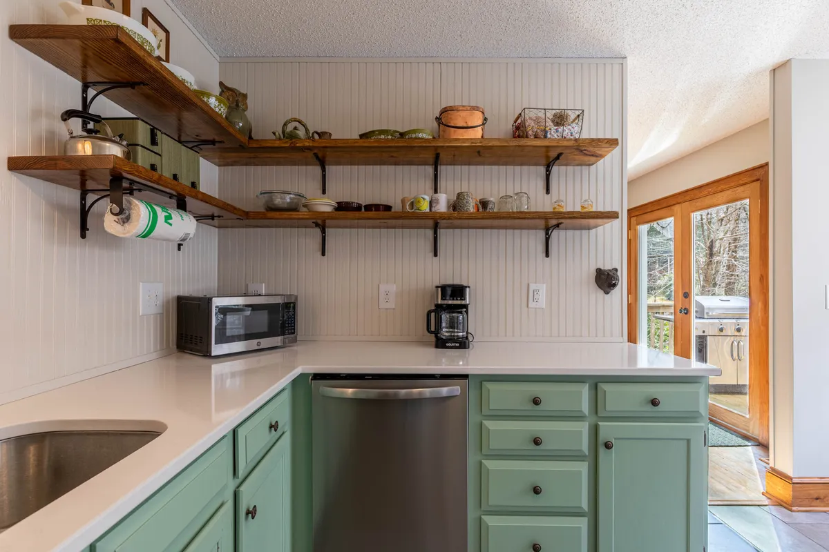 A welcoming kitchen scene with green cabinetry, open wooden shelves, stainless steel appliances, and large windows allowing natural light to flood in. The space is styled with warmth, showing the kitchen as a hub for morning coffee and lively meal prep.