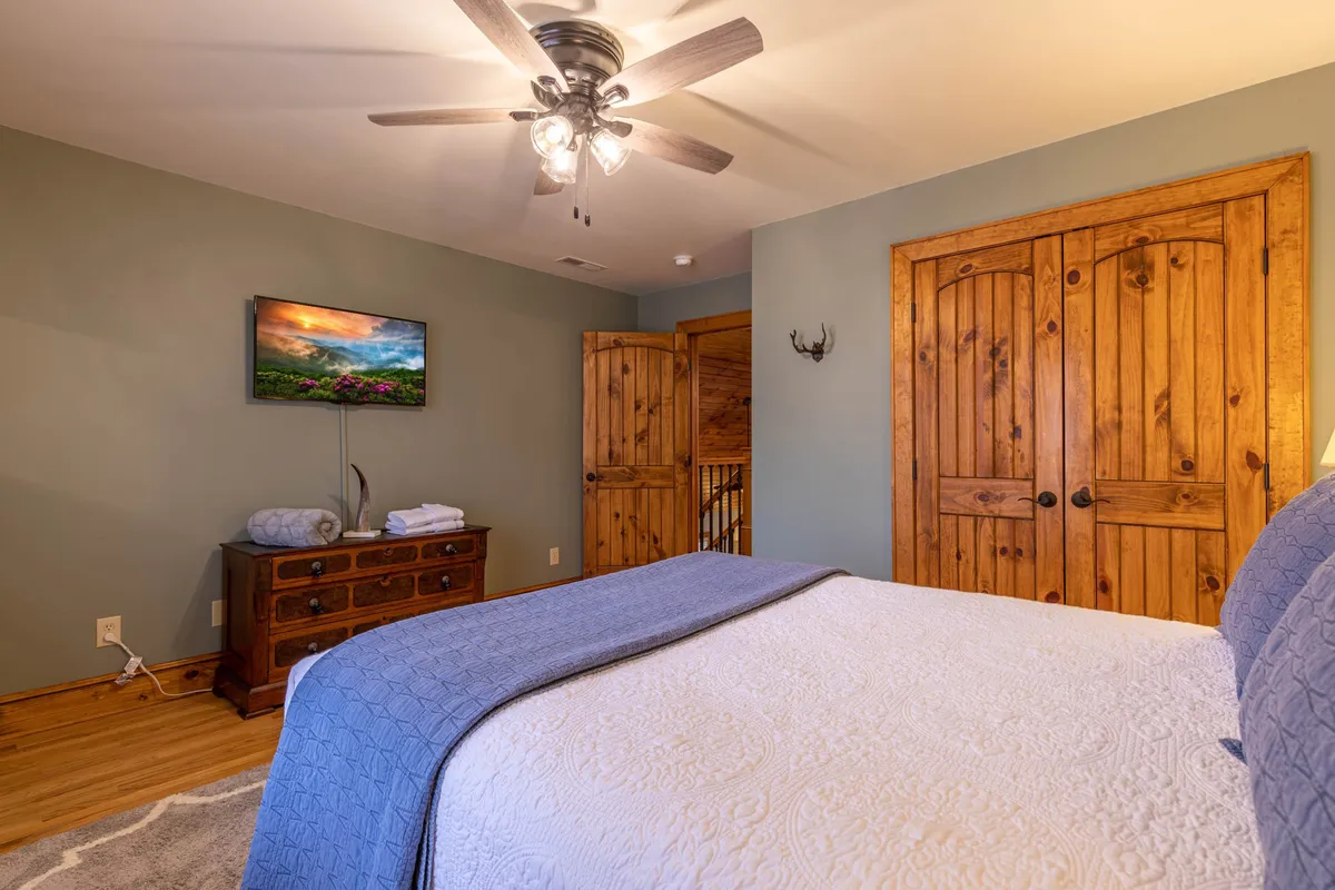 This image features a queen bedroom with a bed adorned with a blue throw blanket, a wooden dresser, wall-mounted TV, and wooden doors. The lighting is warm and the room exudes comfort and relaxation.