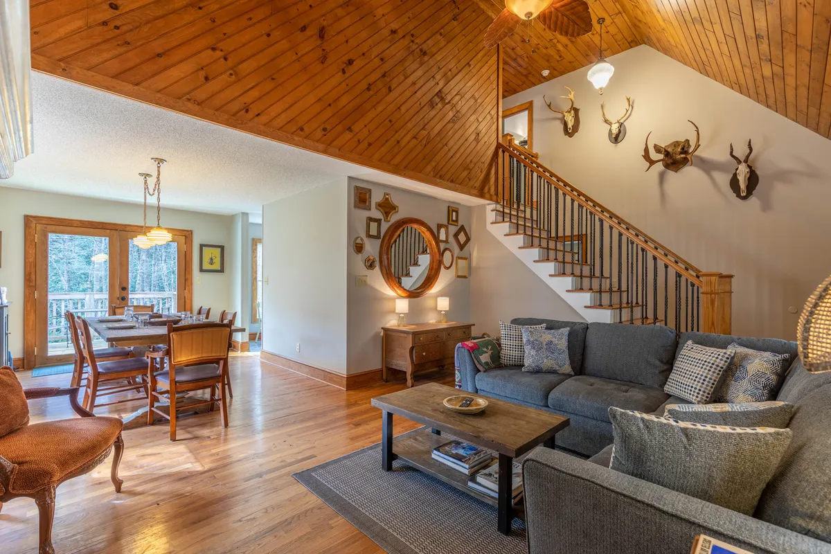 This image presents the main living area of Sleepy Hollow Cabin, featuring a plush sofa, a large wooden dining table, a cozy staircase, and decorative wall mounts and mirrors. The open-plan design is evident, with the sofa and dining area sharing the same bright, inviting space, highlighted by natural light streaming in from large windows. The atmosphere is warm and welcoming, perfect for gatherings and relaxation.