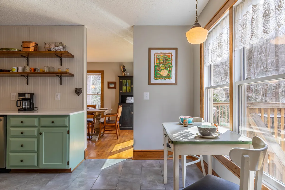 This image captures a small wooden dining table for two nestled by large windows, surrounded by green kitchen cabinets and decorative wall art. The setup is intimate, suggesting a perfect spot for morning coffee or tea while enjoying the view.