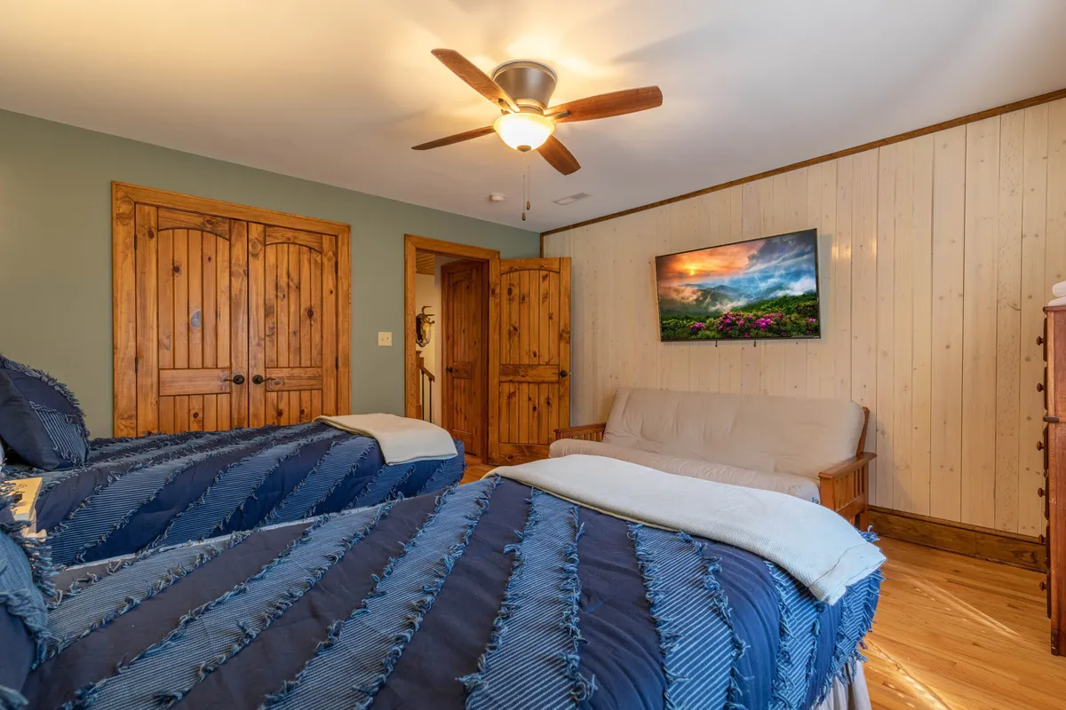 The third bedroom is shown with two twin beds, wooden doors, a TV mounted on the wall, and a sofa. The setting is inviting and ideal for children or additional guests, with cozy bedding and space for relaxation and entertainment.